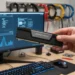 Hand holding a Corsair Vengeance DDR5 RAM stick in front of a computer monitor displaying performance graphs.