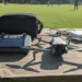 DJI Mini 3 drone and Fly More kit accessories laid out on a weathered wooden picnic table at a sunny neighborhood park with blurred people playing frisbee in the background.