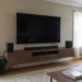 Wall-mounted flat-screen TV above a floating walnut media console with a soundbar, bookshelf speakers, and subwoofer in a naturally lit living room.
