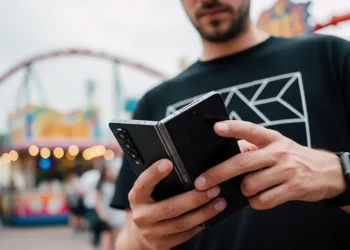 A man holds a dark grey foldable smartphone in a partially folded state at a bright amusement park.