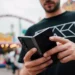 A man holds a dark grey foldable smartphone in a partially folded state at a bright amusement park.