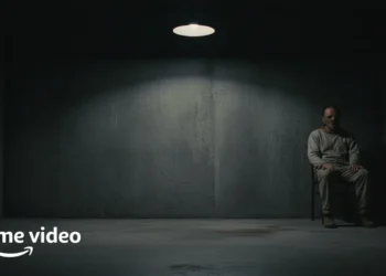 Figure in a restraint mask sits in a dark clinical cell under a spotlight.