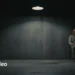 Figure in a restraint mask sits in a dark clinical cell under a spotlight.