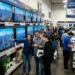 Shoppers comparing prices on new TVs and laptops in a crowded electronics store, illustrating tech trends to watch as gadget prices rise